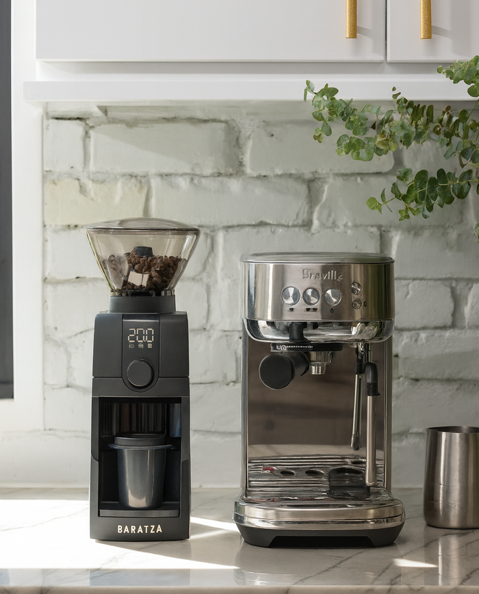 Black coffee grinder and silver espresso machine on a kitchen counter with a white brick wall background.