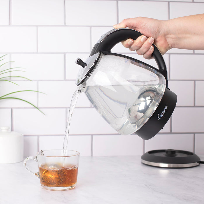 Person pouring water from a Black and polished chrome Capresso 48 oz electric kettle with black handle into a glass mug on a kitchen counter.