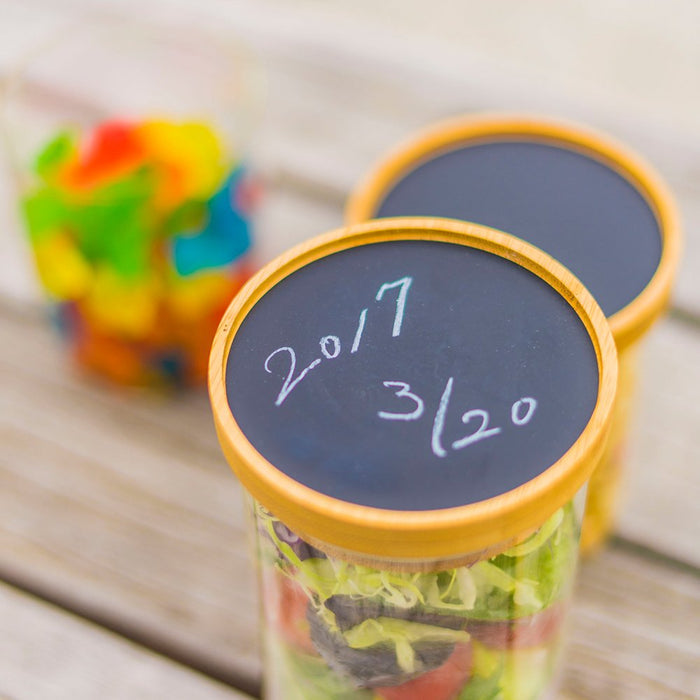 Two jars with black lids displaying '2017 3/20' on a wooden surface, with colorful items in the background.