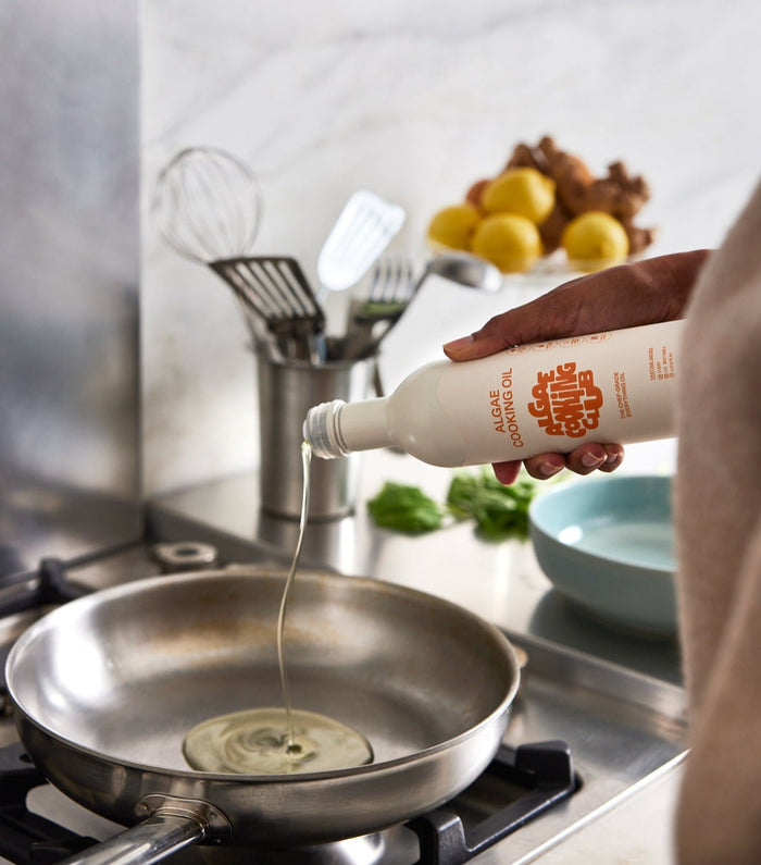 Person pouring cooking oil from a bottle into a frying pan on a stove.
