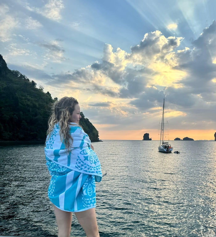 Person wrapped in a teal towel overlooking a serene water body with a sailboat and mountains.