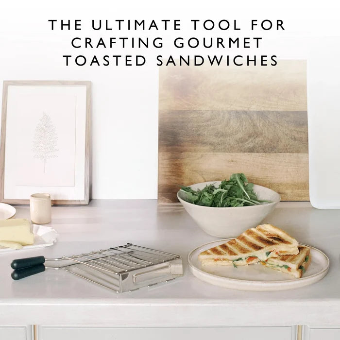 Toasted sandwich on a plate with a bowl of greens, bread, and a sandwich cage on a kitchen counter.