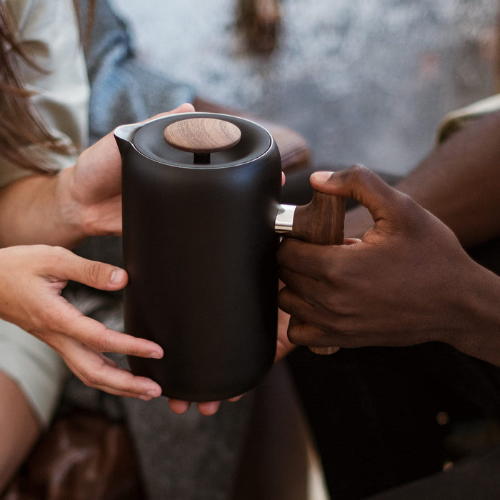 Two hands holding a black french press with wooden lid and handle.
