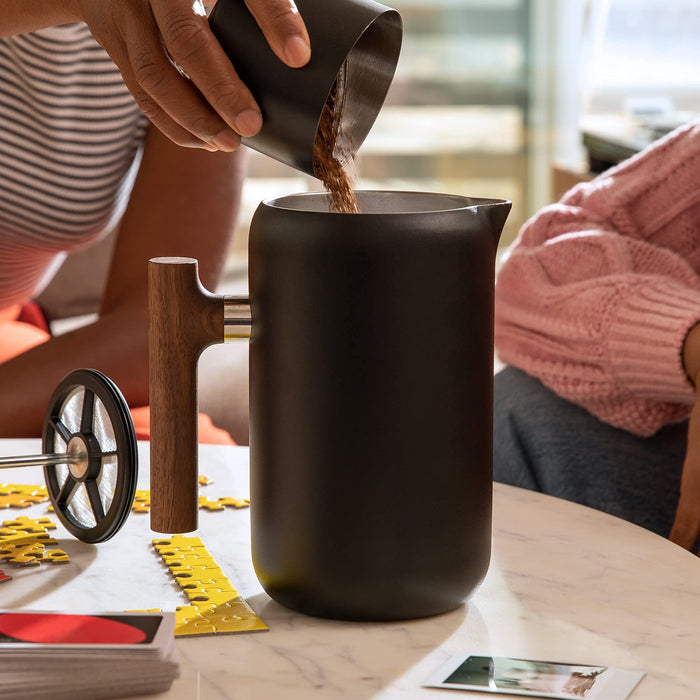 Person pouring coffee grounds into a black french press on a table with a blurred background