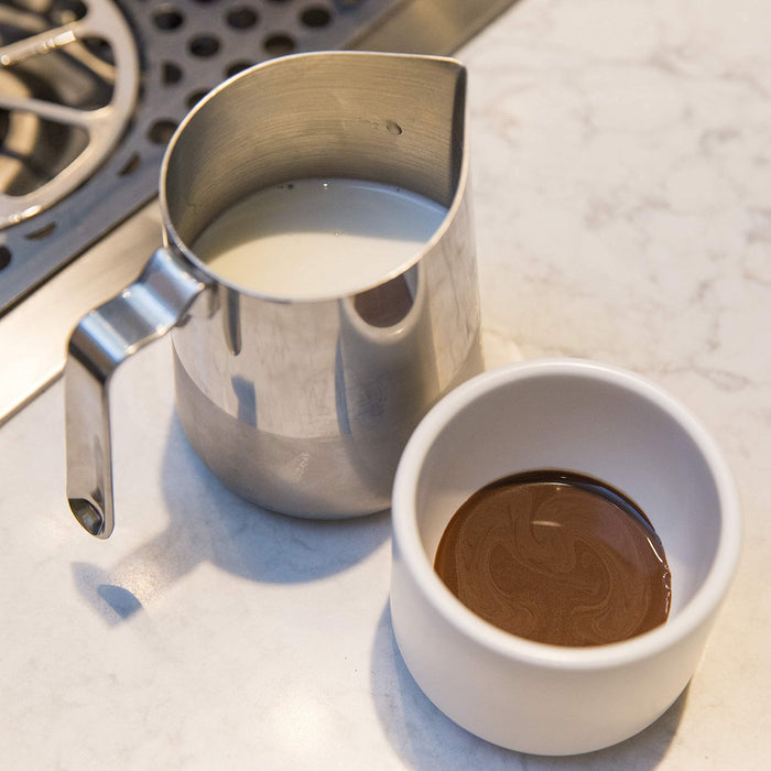 Metal pitcher with milk next to a white cup with coffee on a marble surface