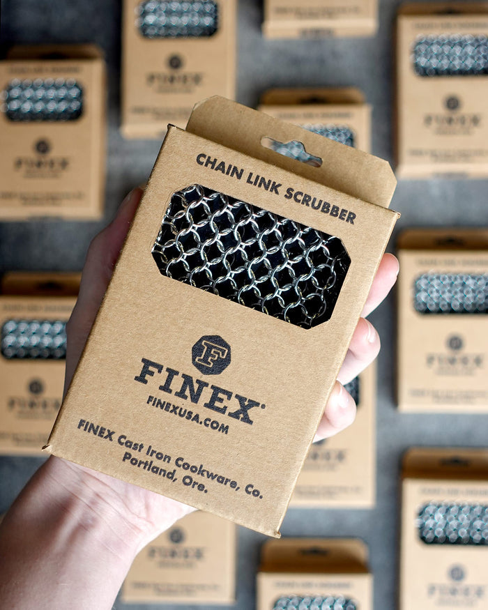 Hand holding a Finex chain link scrubber packaging with multiple boxes in the background.