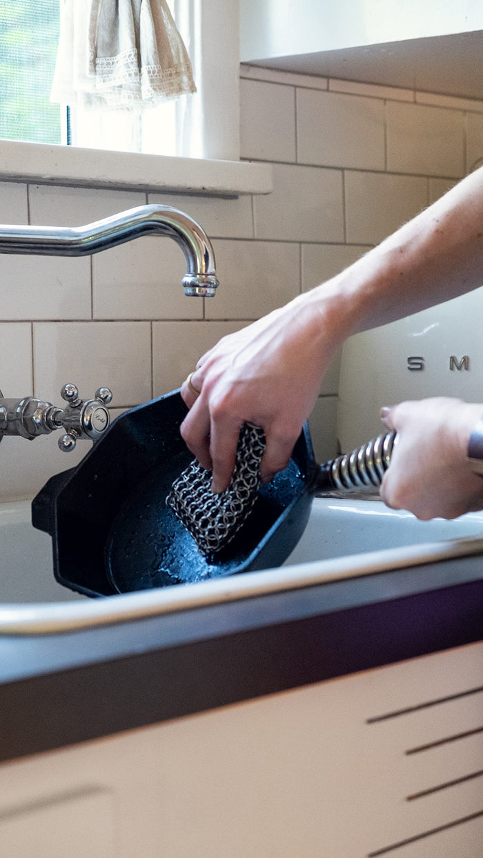 Person cleaning a skillet in a kitchen sink with a chain link scrubber