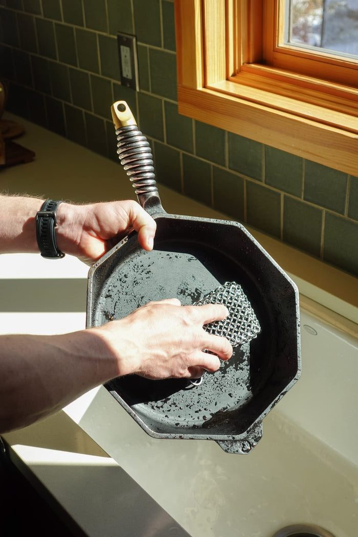 Person scrubbing a cast iron skillet with chain link scrubber in a kitchen.