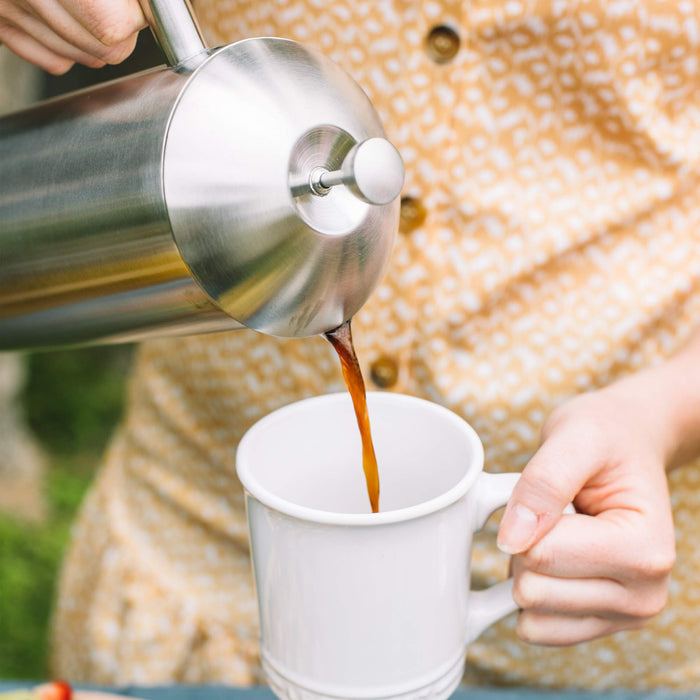 Person pouring coffee from a French press into a white mug outdoors.