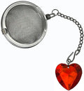 A stainless steel spherical tea infuser with a short chain and a dangling faceted red heart-shaped charm, shown on a plain white background.