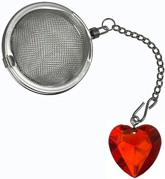 A stainless steel spherical tea infuser with a short chain and a dangling faceted red heart-shaped charm, shown on a plain white background.