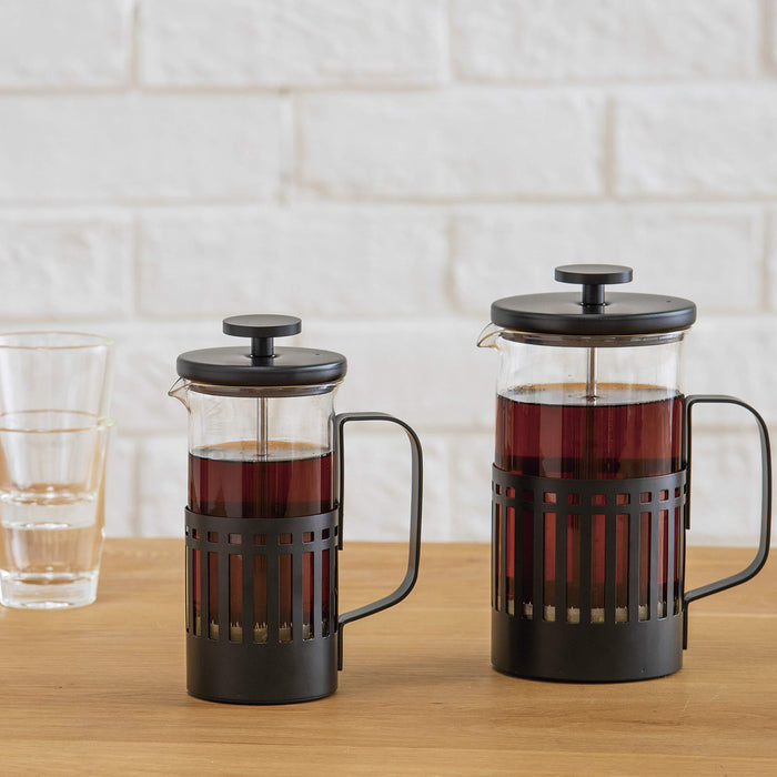 Set of 2 black and clear Hario French press filled with coffee and 2 clear cups in the back displayed on a wooden surface