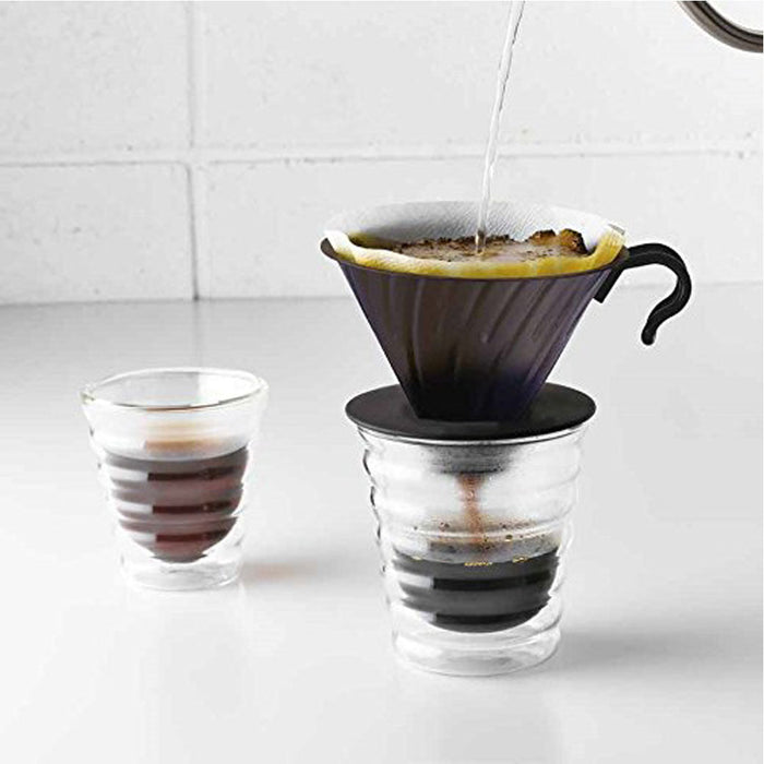 Water being poured into Hario V60 coffee dripper next to a cup of coffee on a white countertop