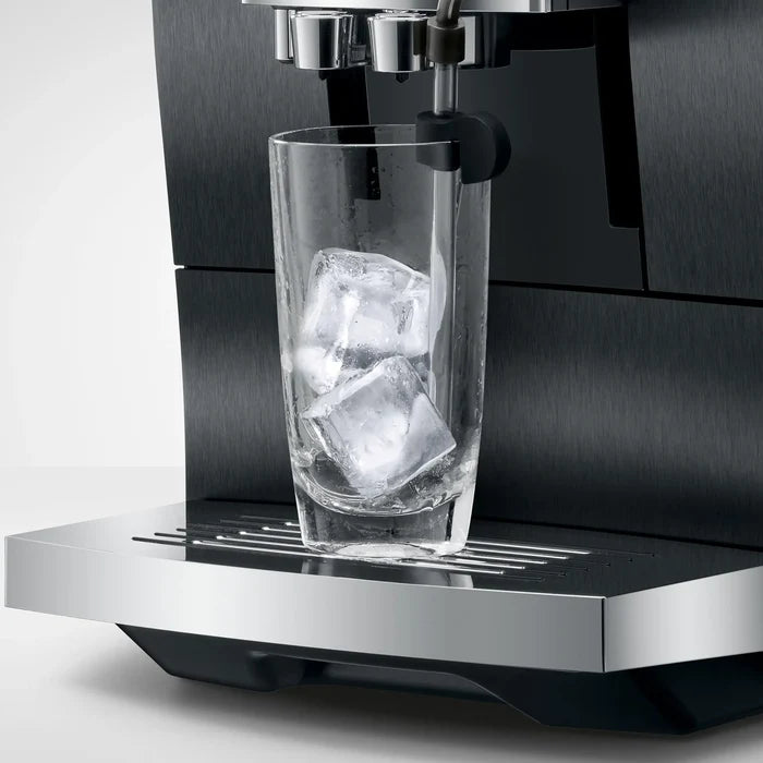 A close-up of a black and stainless steel espresso machine dispensing into a clear glass tumbler filled with ice cubes, set against a clean light gray background.