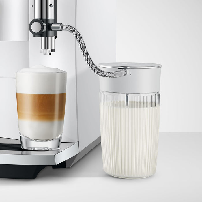 Jura 16.9oz glass milk carafe with lid in attached to a coffee machine with a creamy coffee on a white background