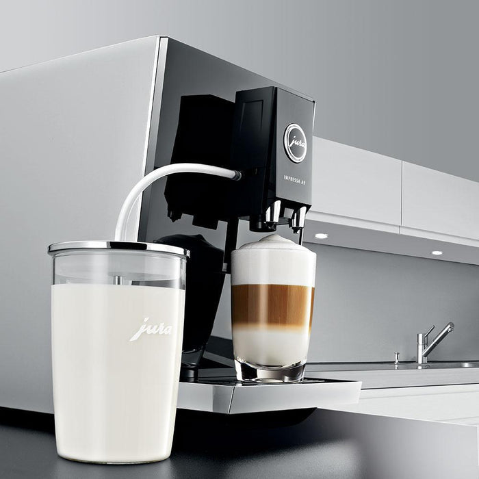.5 liter Jura Glass Milk Container filled with milk and attached to a Jura coffee machine with a large cup of creamy coffee in a kitchen setting