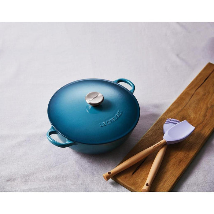 Top view of blue Le Creuset 3.5 Qt. Soup Pot with lid next to board with 2 cooking utensils displayed on a white cloth surface