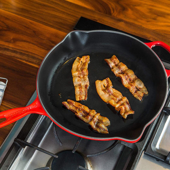 Cerise 6.33" Le Creuset skillet with bacon in it on a stove with wooden countertop background