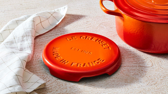 Flame Le Creuset enameled cast iron signature trivet on a wooden background with a kitchen towel and stockpot