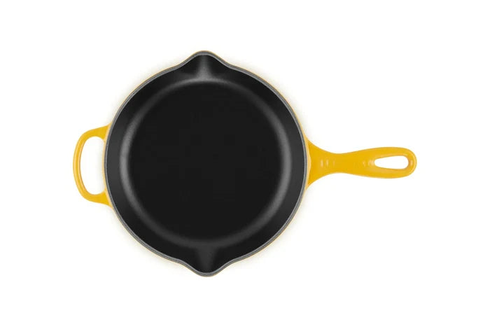 A top-down view of a yellow cast iron skillet with a black non-stick interior, featuring a yellow handle and a small yellow bail on the left side, set against a plain white background.