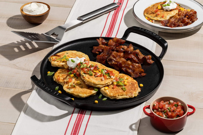 Skillet with pancakes, bacon, and a side dish on a table with a red and white striped tablecloth.