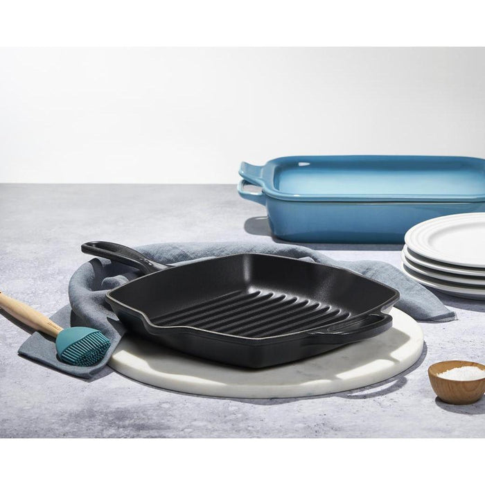 Licorice Le Creuset Enameled Cast Iron Signature 10.25'' Square Skillet Grill with Handle displayed on a marble trivet next to cookware and dinnerware