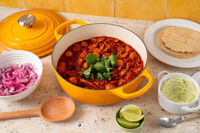 A yellow Le Creuset enameled cast iron pot filled with a red chili stew, surrounded by bowls of onions, lime, avocado, and tortillas on a beige stone countertop.
