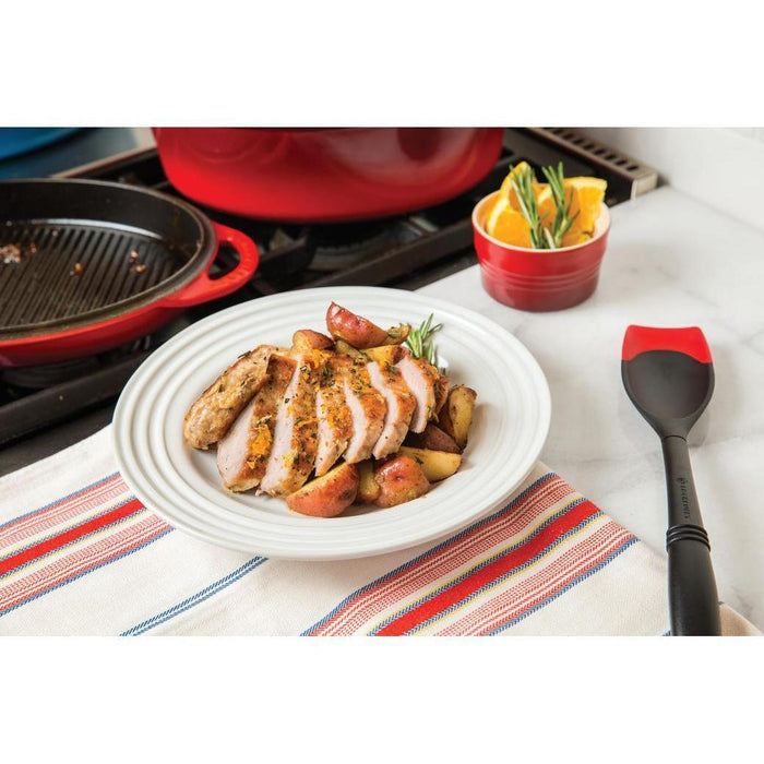 Cerise Le Creuset Revolution Bi-Material Saute Spoon next to a plate of cooked chicken and potatoes on a white countertop