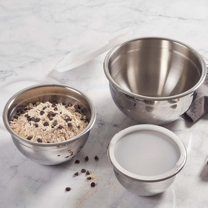 Set of 3 Le Creuset stainless steel mixing bowls with lids with cookie mixture inside medium sized bowl placed on a marble surface
