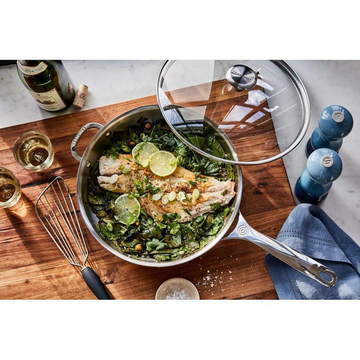 Top view of 12 inch Le Creuset Signature Tempered Glass Lid and skillet with cooked food displayed on a wooden cutting board next to cooking materials