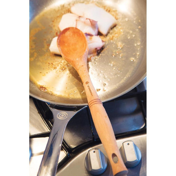 Le Creuset 10″ Stainless Steel Fry Pan with fish cooking and a wooden spoon displayed on a stove top
