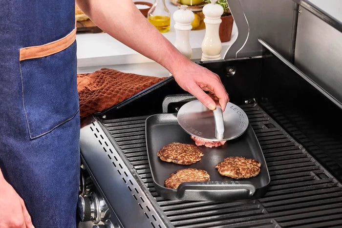 A person wearing a blue apron uses a round metal grill press with handle to press a cooked burger patty from a black square grill pan on a gas grill.