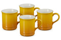 Four Le Creuset ceramic mugs in a warm mustard yellow color with a glossy finish and white interiors, arranged on a clean white background.
