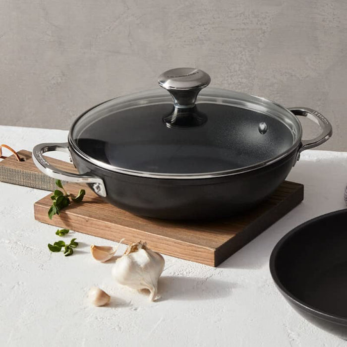 Black frying pan with a lid on a wooden cutting board against a gray background