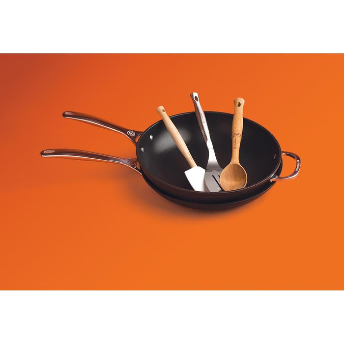 Le Creuset Toughened Nonstick Stir Fry Pan, 12″ with cooking utensils displayed on an orange background