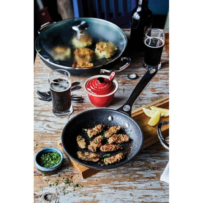 Le Creuset Tri-Ply Stainless Steel Nonstick Fry Pan with fried food and cooking materials in the background