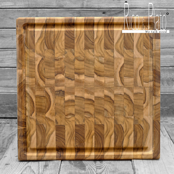 A square, brown teak wood cutting board with juice grooves and handles, crafted from durable end grain construction on a wooden background