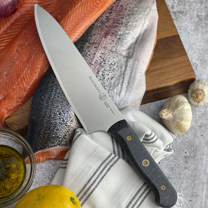 8 inch chef knife next to sauce, salmon, garlic, and napkin on a cutting board placed on a grey surface