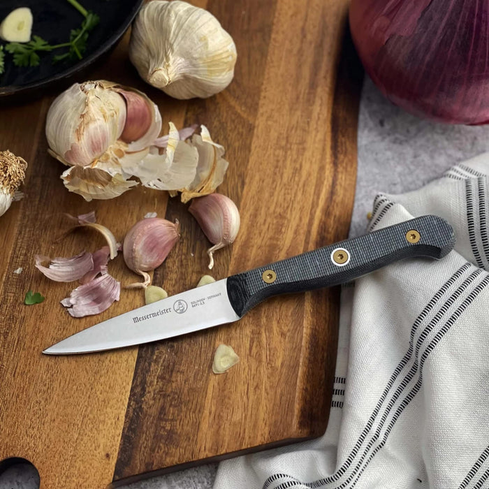 3.5 inch paring knife next to garlic and onion on a cutting board displayed on a grey surface