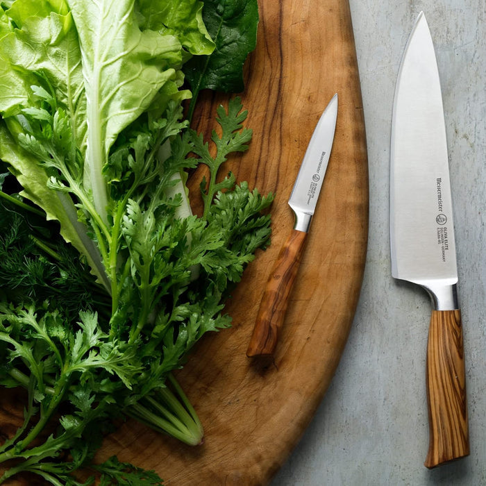 A chef knife and a pairing knife with wooden handles on a wooden cutting board next to fresh green leafy vegetables.