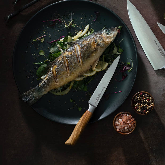A few knifes next to a plate of a cooked fish and vegetables next to 2 small bowls of seasonings on a brown surface
