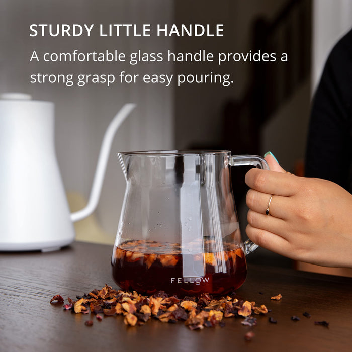 Glass coffee pot with handle being held, surrounded by spices on a wooden surface.
