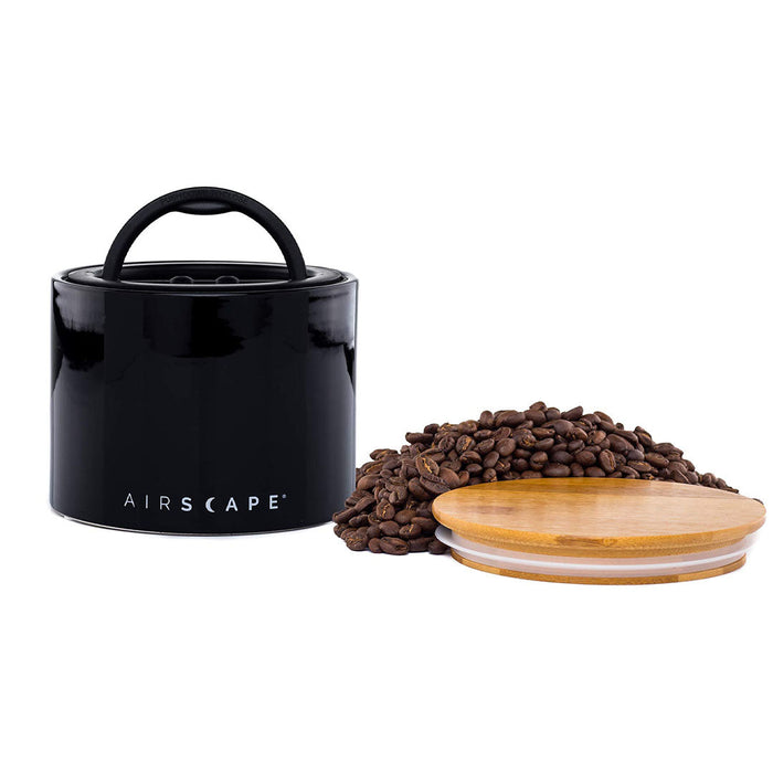 Black air tight container with black handle and lid, with coffee beans on a white background.