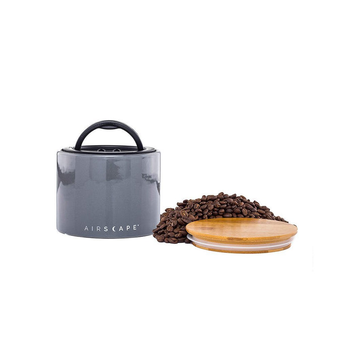 Grey air tight container with black handle and lid, with coffee beans on a white background.