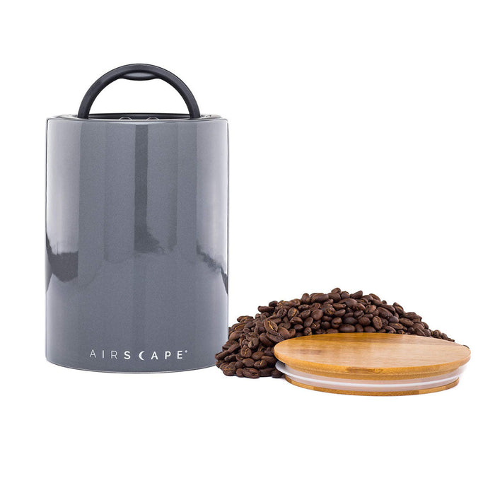 Grey air tight container with black handle and lid, with coffee beans on a white background.