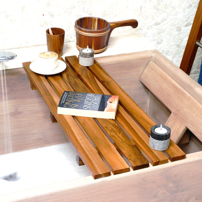 Teak tub caddy with horizontal planks on a tub with bathroom essentials on top of tub caddy