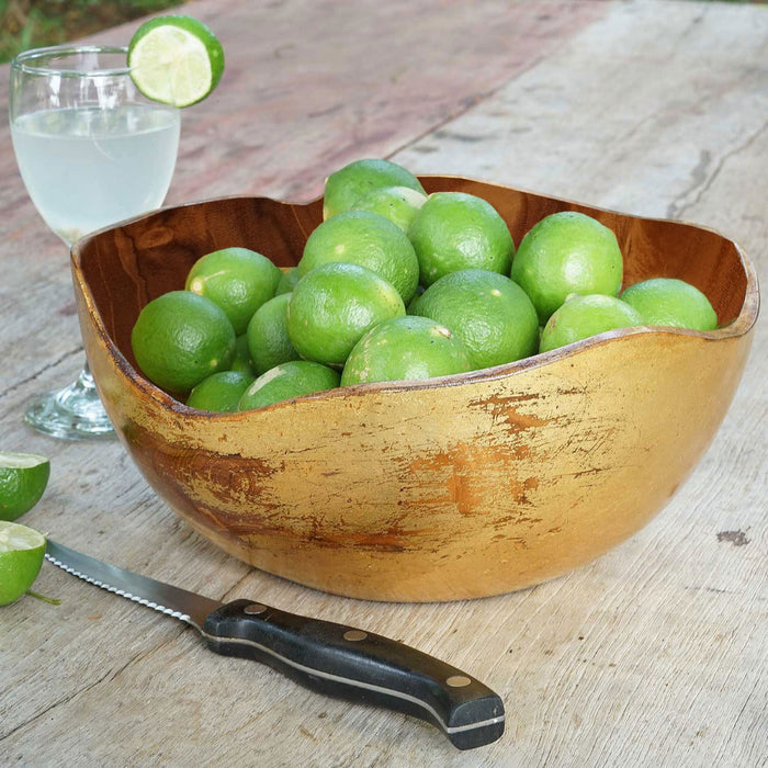 Extra large teak bowl with distressed gold leaf exterior with limes placed inside next to knife and glass of lime water displayed on a wooden surface
