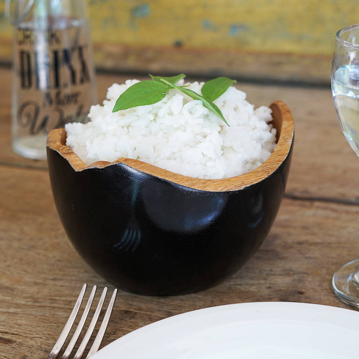 A hand-painted teak bowl with a deep black exterior and natural wood grain interior, containing white rice garnished with a green leaf.