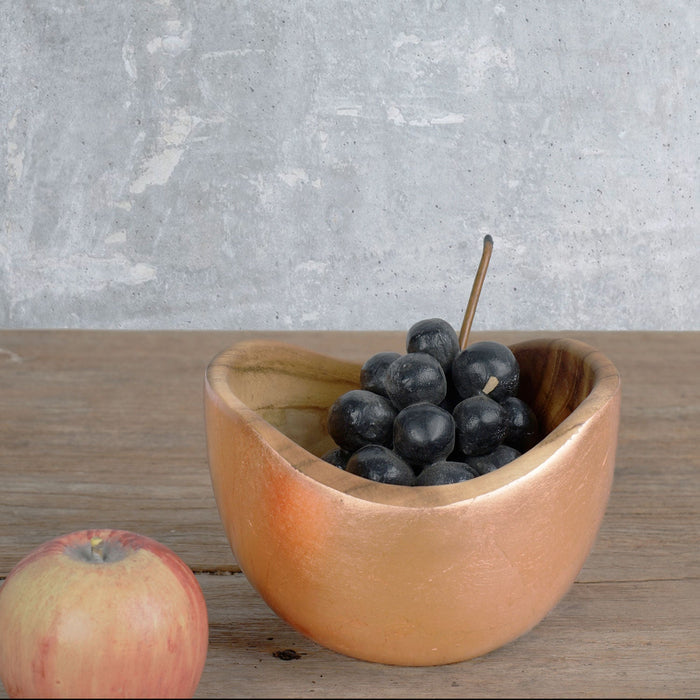 Teak bowl with copper leaf exterior with waxed grapes inside displayed next to a waxed apple on a wooden surface
