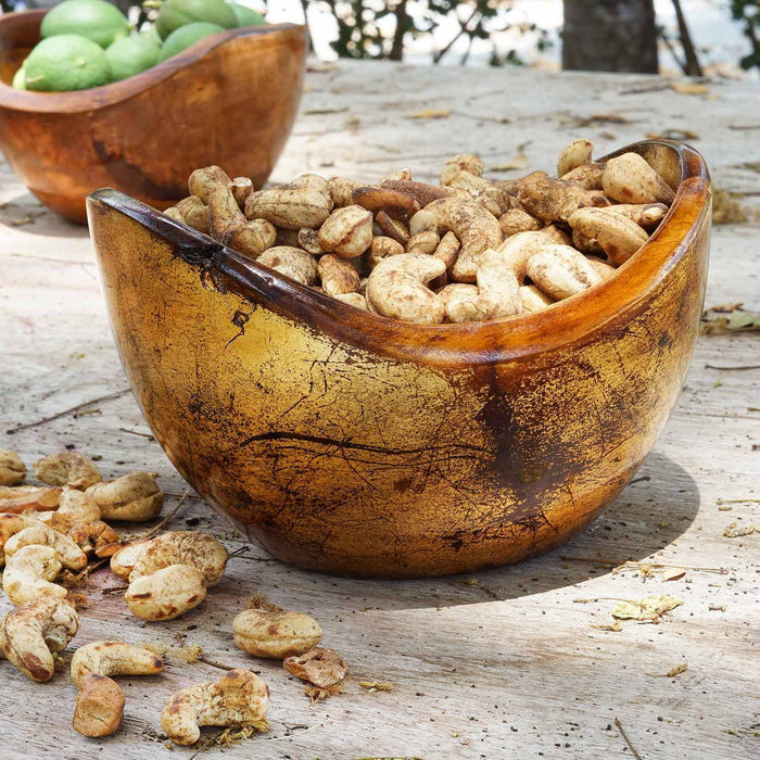 2 Rico & Plato adagio teak bowl with distressed copper leaf exterior one in the back with limes inside and the other in front with cashews inside placed on a wooden surface next to cashews